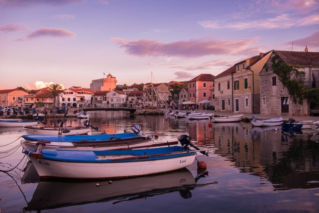 Vrboska village at sunset: the sky is lit in purple and there are small boats in the canal, surrounded by stone houses on each side.