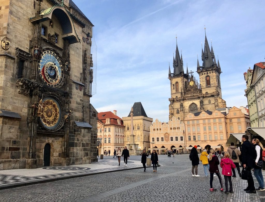 A view of Prague's Astronomical Clock on the left, with a blue and gold clock and several rings moving around. In the background, Old Town Square with pointy church steeples sticking above the peach-colored buildings.