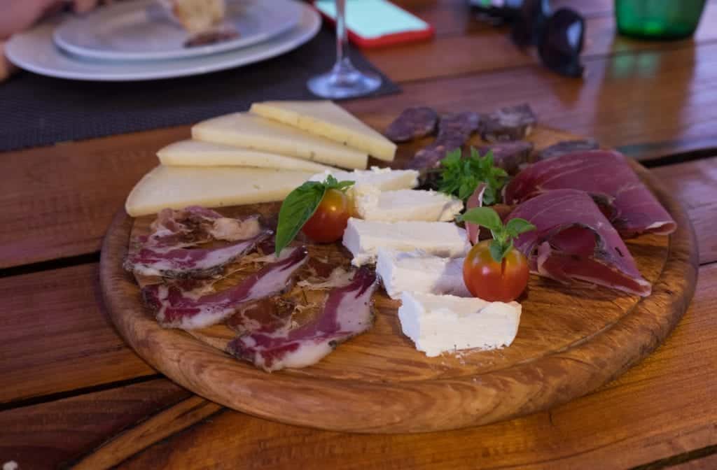 A wooden platter topped with cured meats, two kinds of cheeses, and two cherry tomatoes.