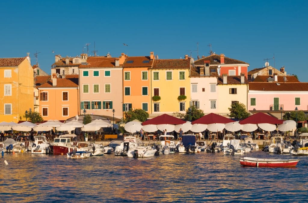 Cafes along the waterfront in Rovinj underneath umbrellas; bright yellow buildings with orange roofs behind them.