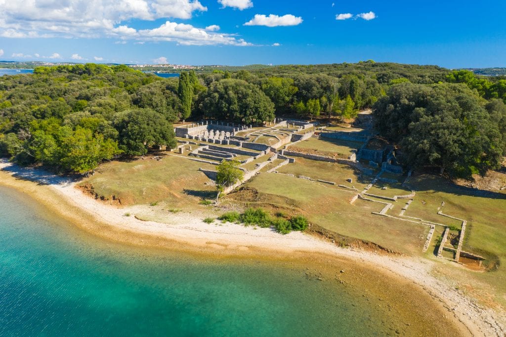 View over the Brijuni Islands: green lush forests and stone walls on land, and a white pebbly beach leaning into deep green water.