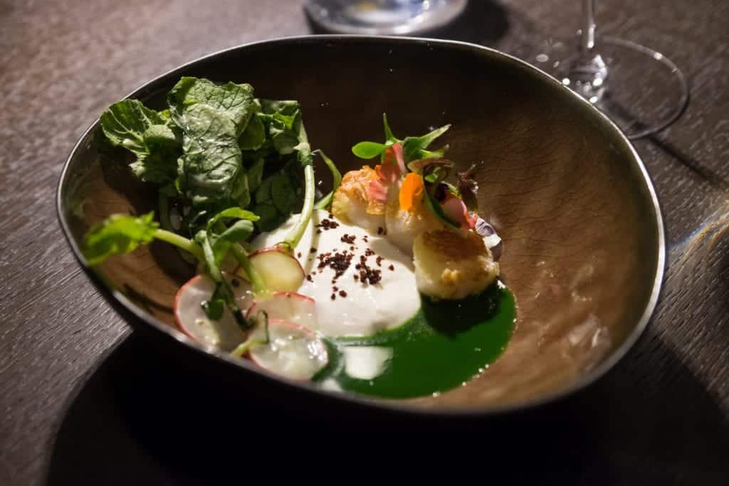 A brown ceramic bowl filled with three fried scallops and some greens, horseradish sauce, radishes, and green sauce.