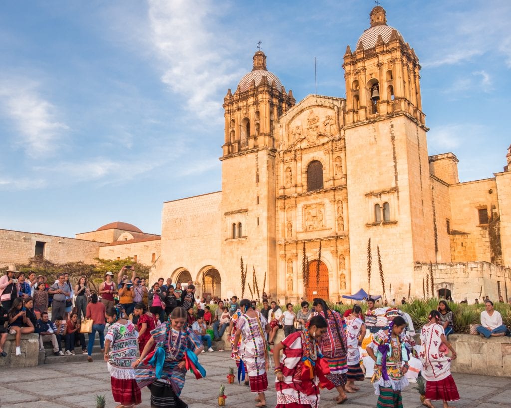 Indigenous women dancing in brightly colored dresses in front of the brightly lit cathedral underneath a blue sky in Oaxaca, Mexico.