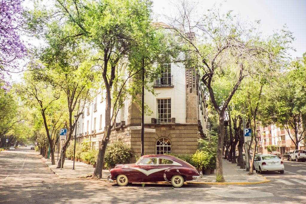 An old-fashioned maroon car parked in front of a white building with two streets on each side of it, lined with jacaranda trees.