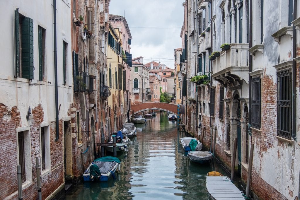 A canal in Venice surrounded by brick-and-plaster homes with balconies brimming with flowers, a bridge in the distance.