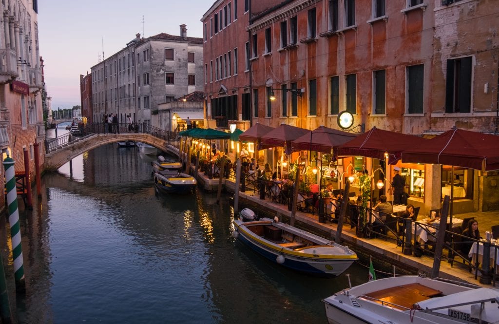 People sitting at a restaurant lit by golden street lamps on the edge of a canal, small wooden boats in the canal.