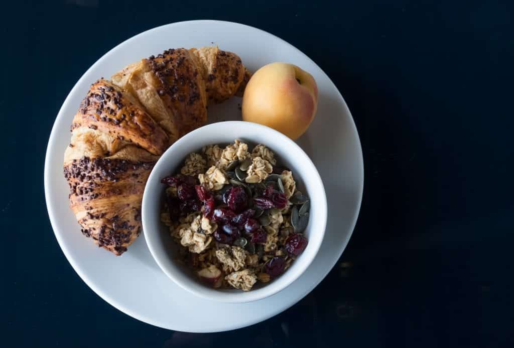 A plate topped with a croissant, apricot, and bowl of granola.