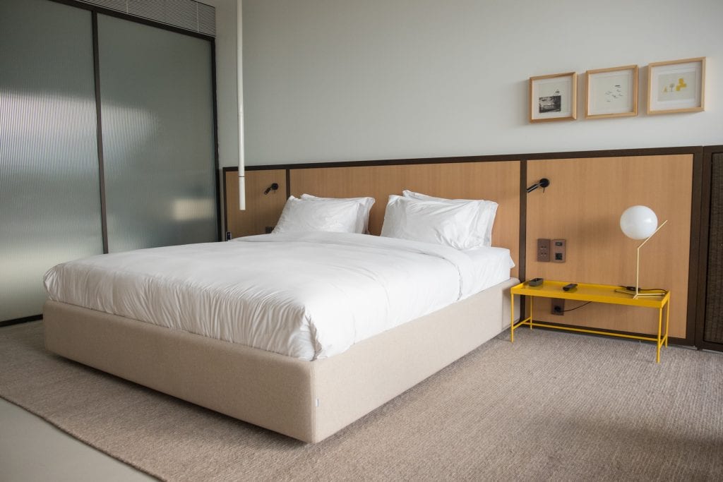 A simple room with a big white bed, yellow metal side table, and three natural wood framed prints on the wall.