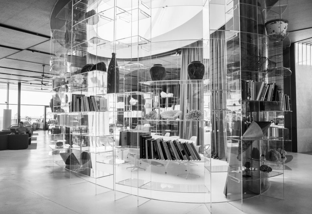 An office area in the Grand Park Hotel: a clear oval-shaped room lined with shelves topped with books and vases, a long table surrounded by chairs in the room.