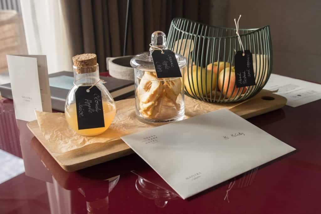 Gifts at the hotel: an envelope reading "Ms. McCulley" plus a container of cloudy apple juice, a clear jar filled with apple rings, and a wire fruit basket filled with apples.