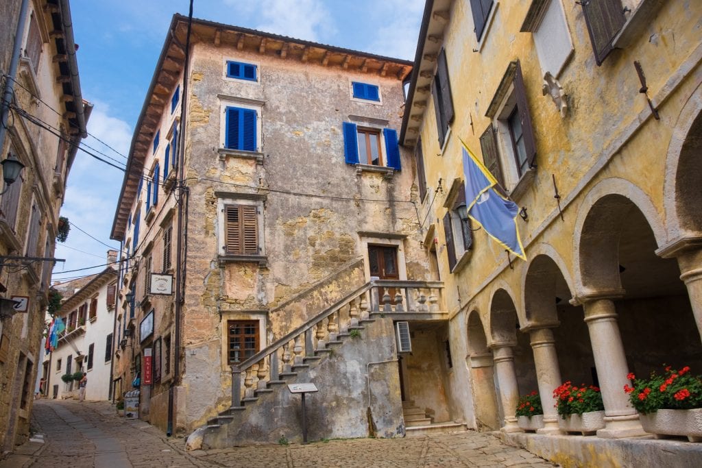 The stone city of Grožnjan, Croatia, with a courtyard, some buildings with pillars, one building with bright blue shutters.