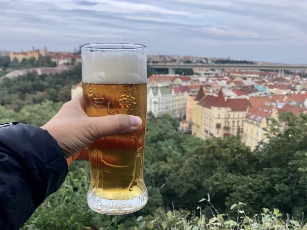 Kate's hand holds a beer while overlooking the treetops and yellow and orange buildings of Prague