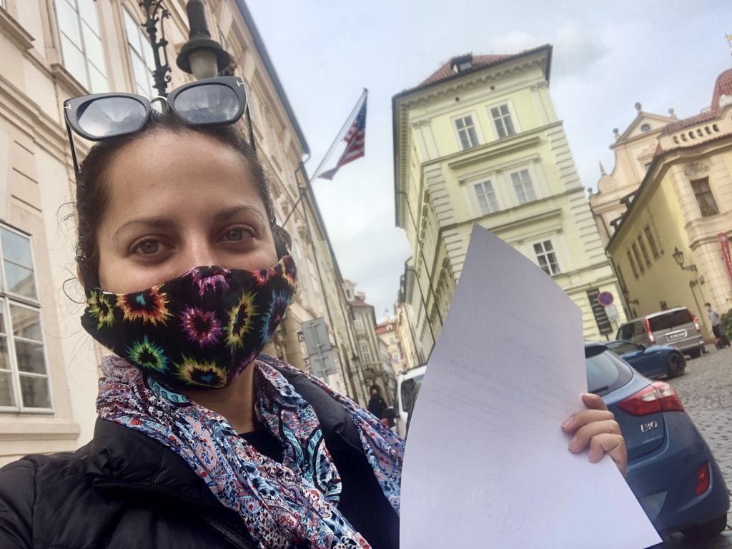 Kate wears a mask and stands in front of the US Embassy in Prague holding a piece of paper that was notarized