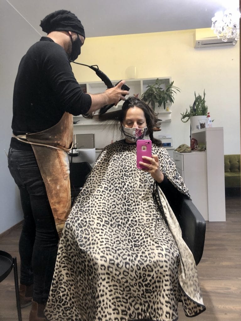 Mirror selfie of Kate getting a haircut in a leopard-print bib, wearing a mask. Her stylish wears a mask while blow-drying her hair.