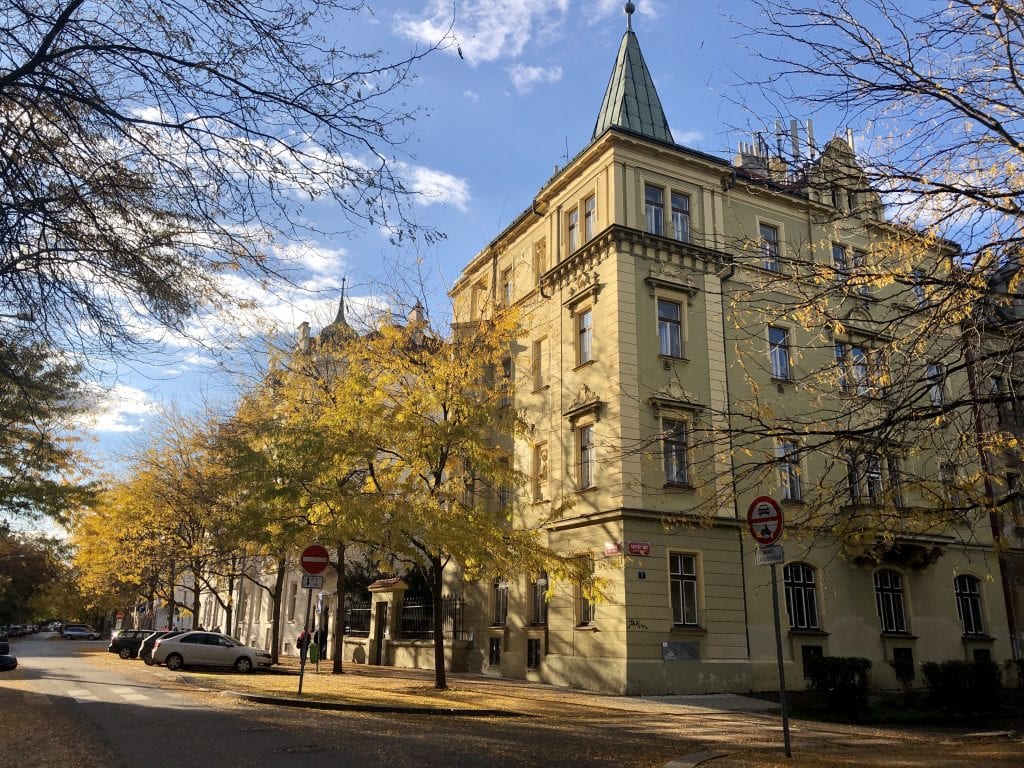 In Karlín: a green crenellated building next to several trees with yellow leaves underneath a blue sky.