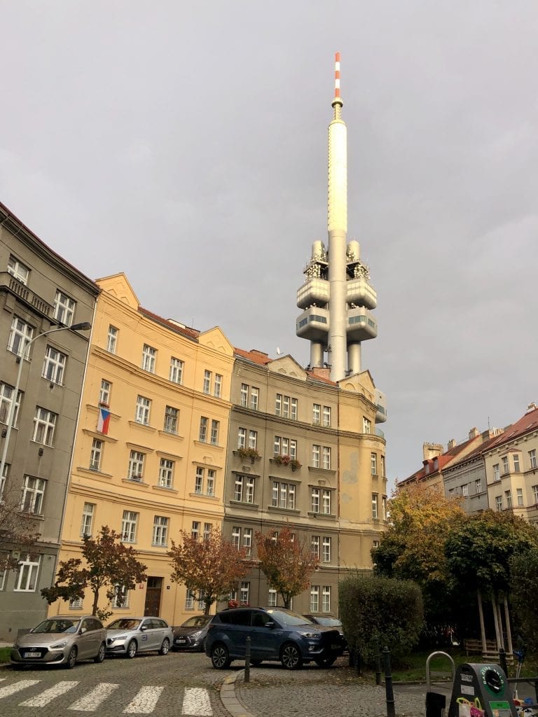 Yellow and brown buildings in the foreground, one draping a Czech flag from the window, and in the background, the silver, space station-like TV tower.