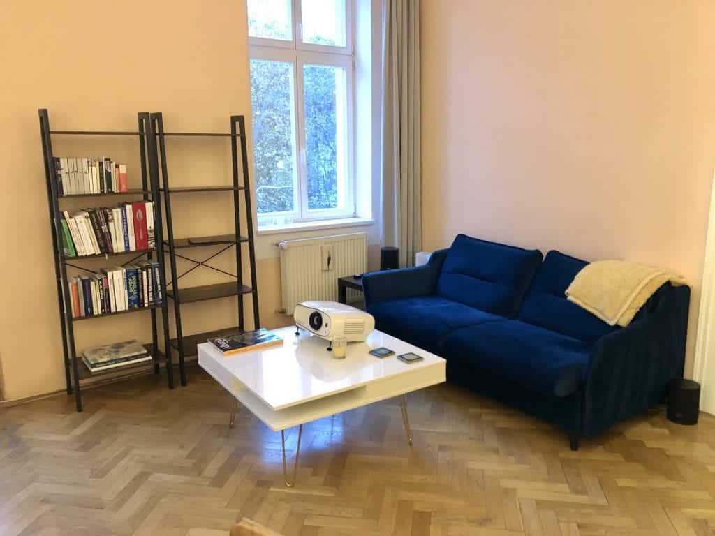 The new living room: two bookcases with industrial black metal and dark oak wood shelves, a white glossy coffee table with brass hairpin legs, and a modern teal sofa with a yellow blanket on top.