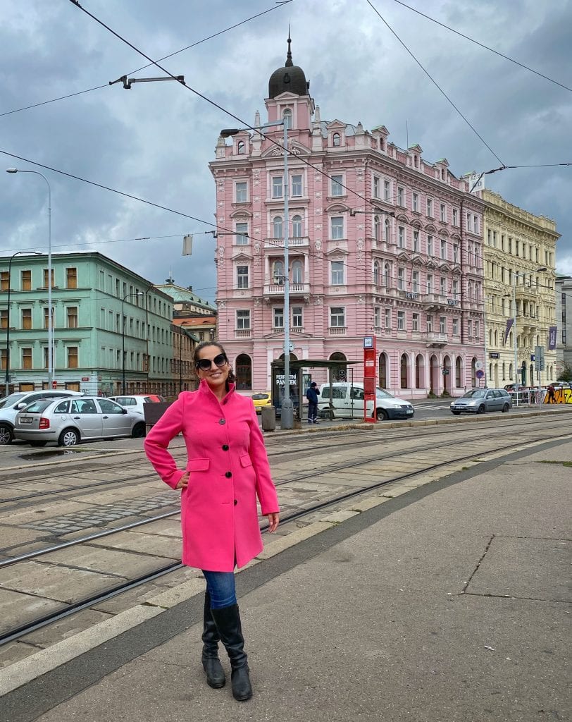 Kate smiling in a hot pink peacoat that hits just above her knees with an upward facing collar. She also wears jeans and tall black boots and big black sunglasses. She is smiling and in the background is a two-tone pink crenellated building.