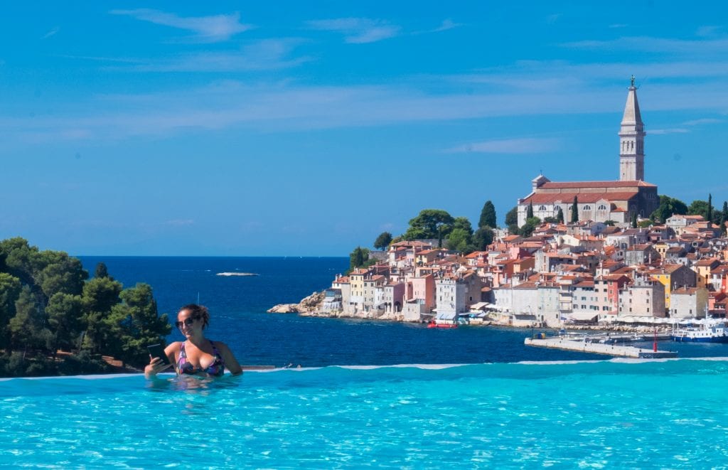 Kate sits in a sky-blue infinity pool, reading her Kindle, the skyline of Rovinj in the background, complete with its church steeple sticking above everything else, under a bright blue sky.