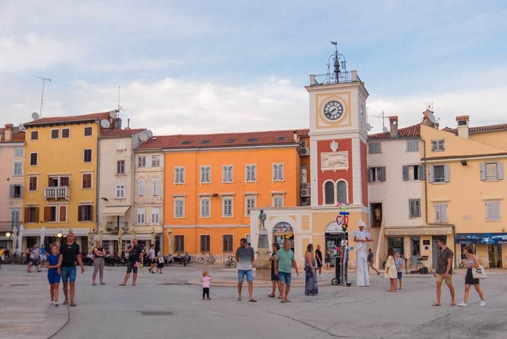 A town square with a yellow, red, and white clock tower, and street performers performing to a crowd.