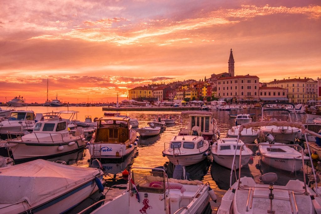 A stunning orange-yellow-pink sunset with the tower of Rovinj in the background; in the foreground are small white boats.