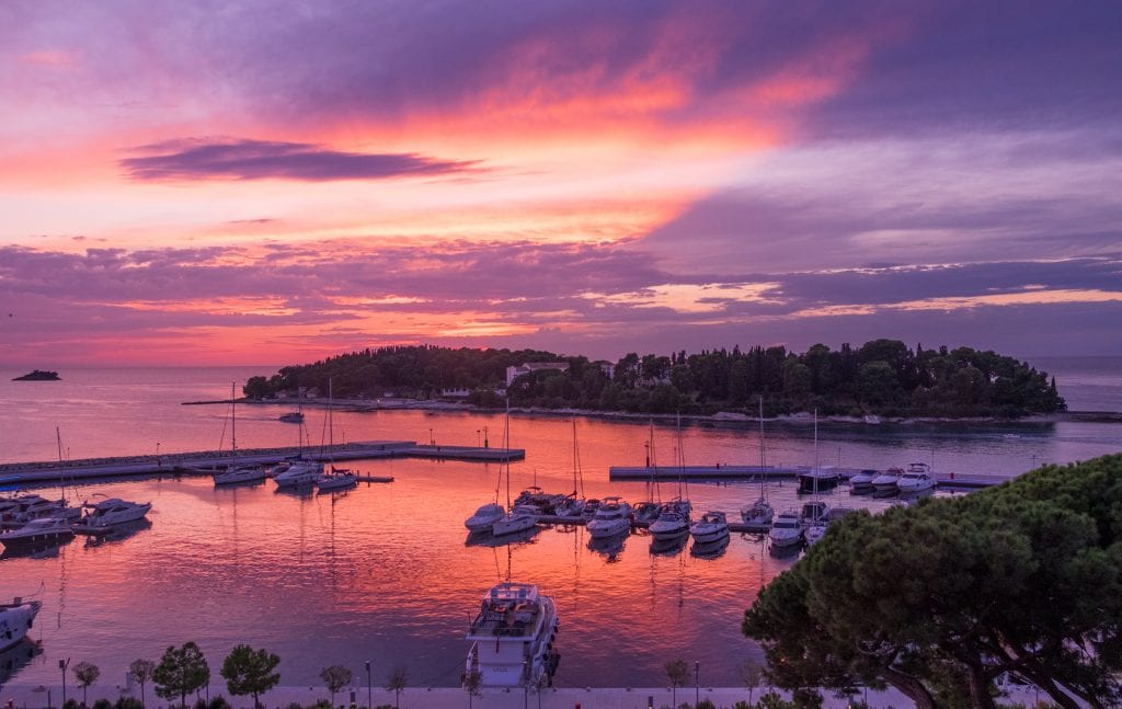 A purple and pink sunset behind a small green island; in the foreground, boats docked at piers.