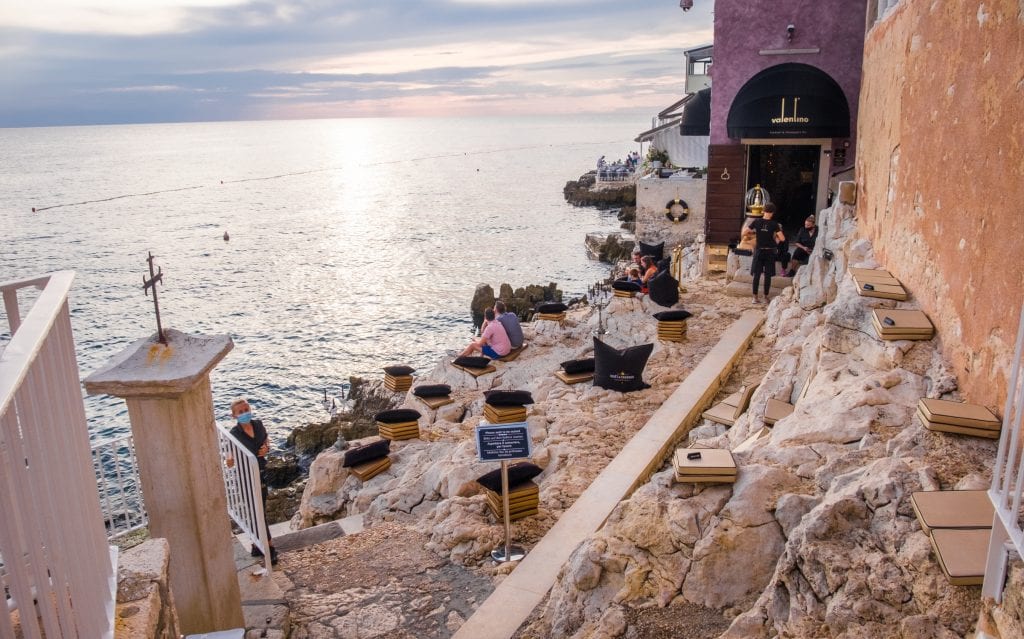 Valentino Bar: a rocky outside seating area topped with seat cushions, all on the edge of the ocean. Waiters wear face masks and serve drinks. A cloudy gray sky.