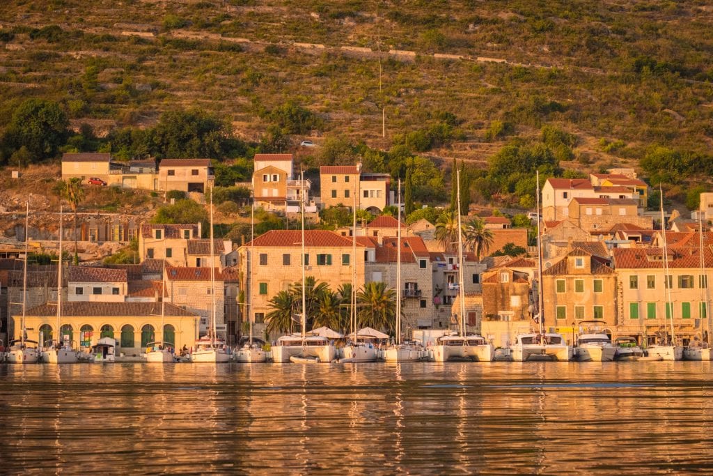 Sunrise in Vis: a view across the water, white sailboats lined up on the shore, sun-drenched brick buildings and hills in the background.
