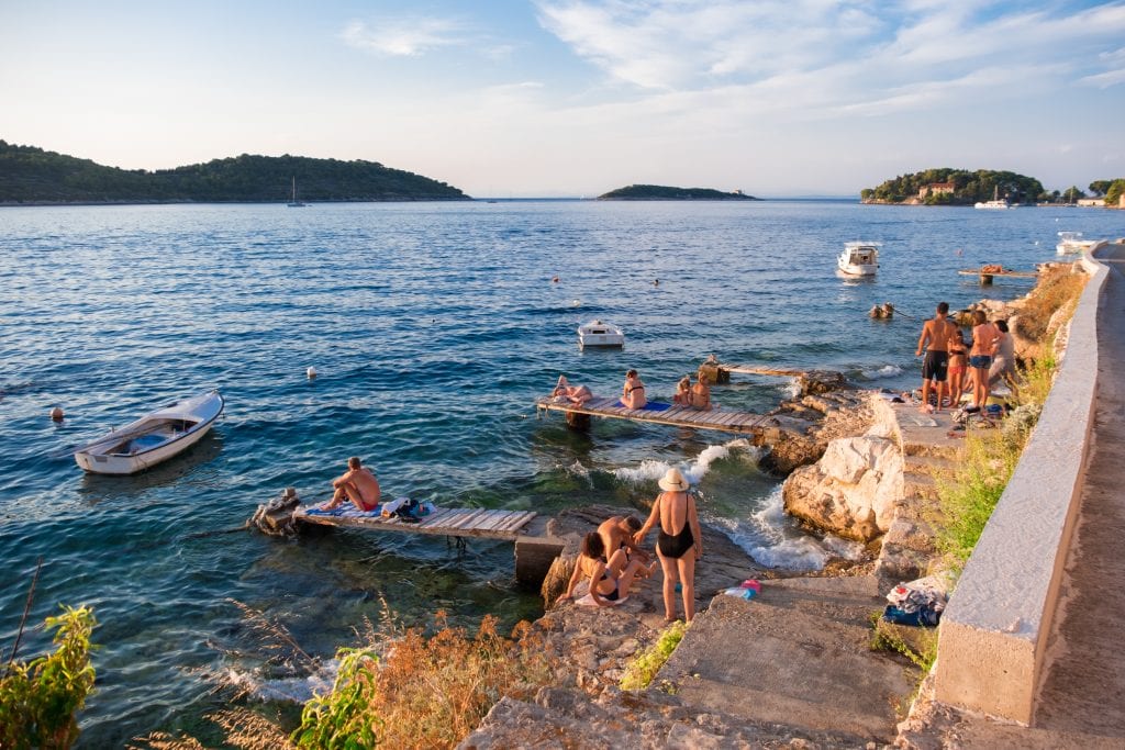 People of various ages hanging out on wooden docks leading into the water, small boats docked close by.