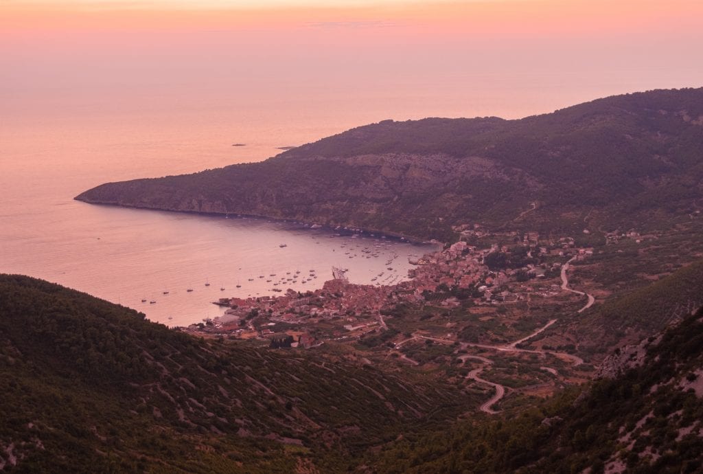 A pink sunset from the top of Hum Mountain, overlooking mountaintops and the town of Komiza in front of the sea below.