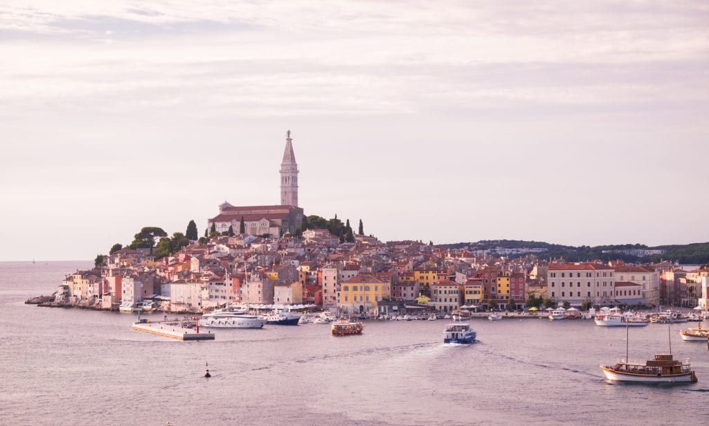 The Rovinj city skyline just before sunset: the same skyline with the warm-colored buildings along the water's edge with a church and steeple sticking straight up: the entire photo has a mild purple near-sunset tone.