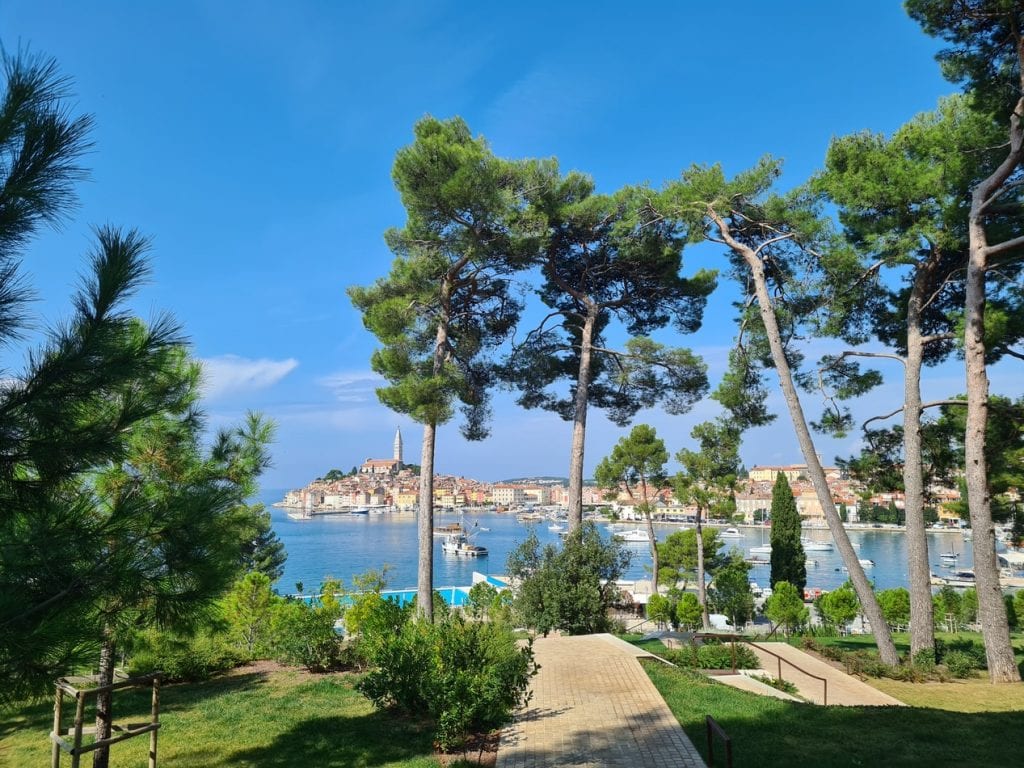 The city of Rovinj in the far distance, as seen from a green island with lots of pine trees partially obscuring the view.