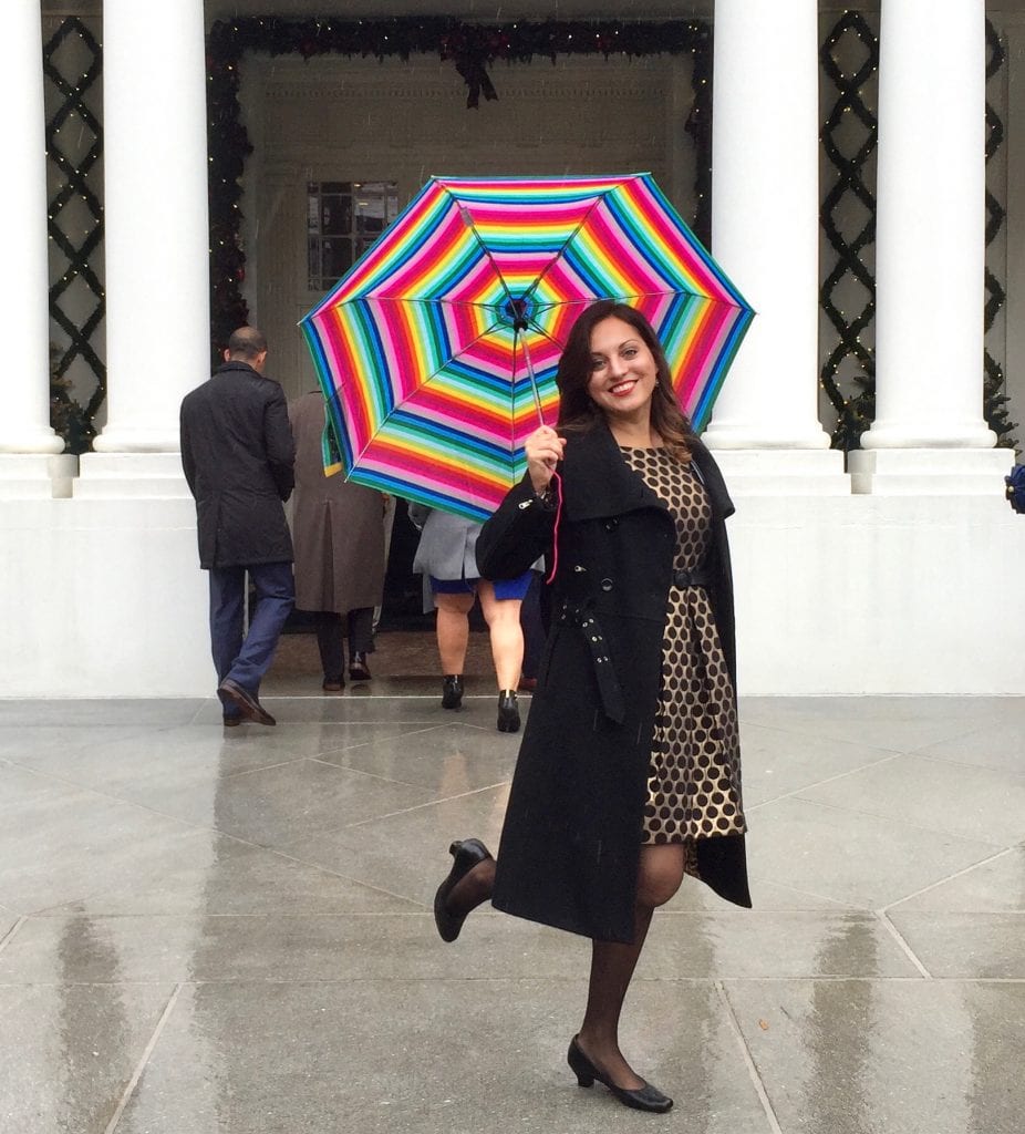Kate wears a black and gold polka dot dress and long black peacoat, holding a rainbow striped umbrella, and smiling with one leg perched up behind her, standing in front of a White House entrance.