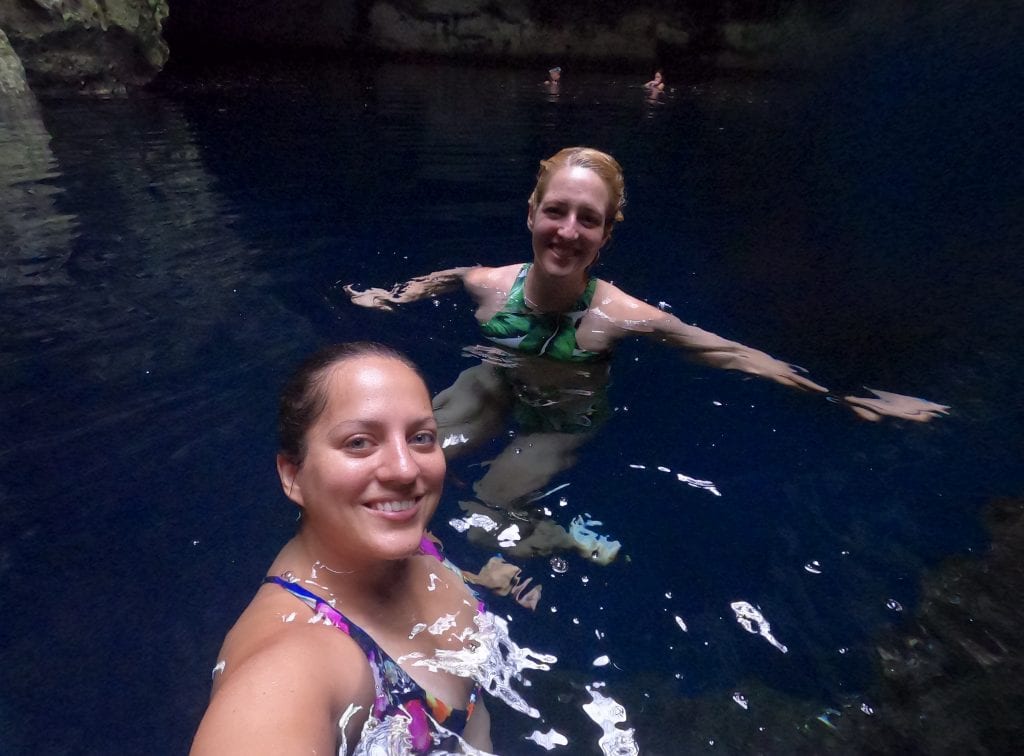 Kate and Sarah float in the clear water of a cenote, surrounded by cave walls.