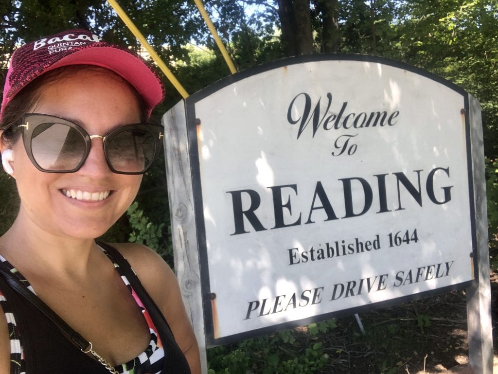 Kate wears sunglasses and her Bacalar baseball cap and poses in front of an outdoor sign that reads "Welcome to Reading. Established 1644. Please drive safely."