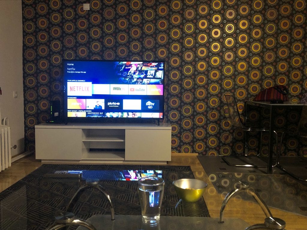 View from the couch in Novi Sad: a glass coffee table, a TV with Netflix pulled up against the wall. The wall is covered in a wild brown, gold and green 70s disco print wallpaper.