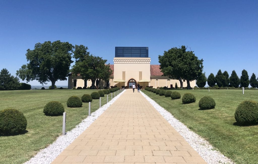 A fancy-looking winery, looking like it's almost out of California, and a stone path leading to it, surrounded by small round manicured bushes.