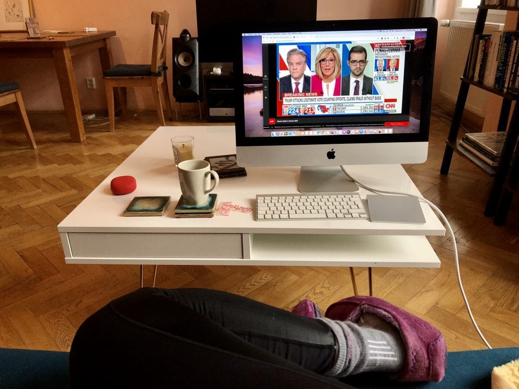 The white coffee table covered with a desktop computer and keyboard, cup of tea, kindle, candle, and packs of pepto-bismol tablets. You can see Kate's legs on the couch clad in leggings, merino wool socks, and suede slippers.