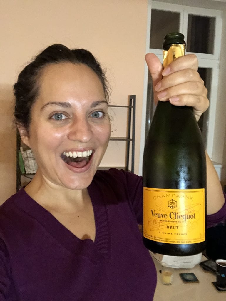 Kate holds a bottle of Veuve Clicquot champagne in her hand and has a huge smile with her mouth open.