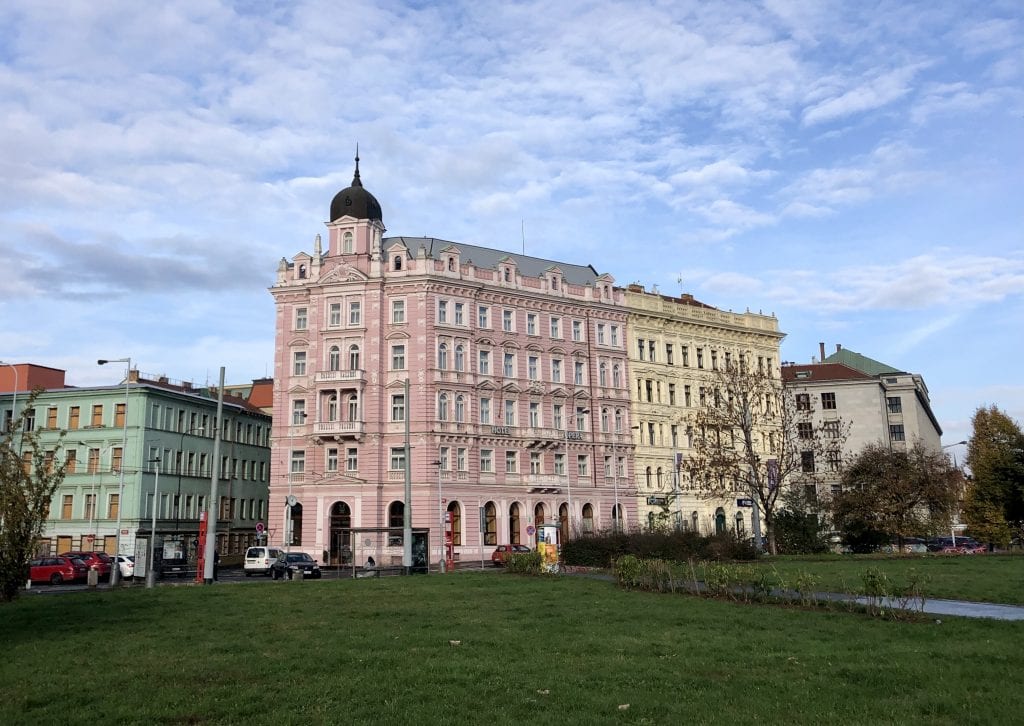 A two-tone pink crenellated building in front of a lawn in Prague, underneath a blue partly cloudy sky.