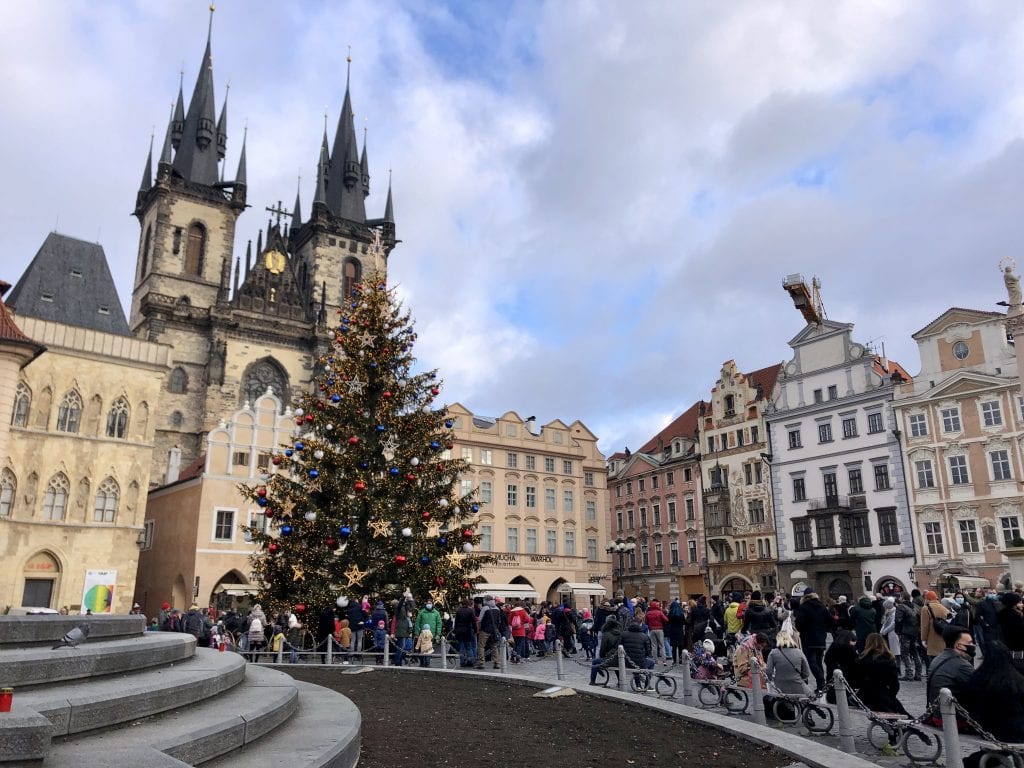 Prague's Old Town Square, with its multi-colored crenellated buildings and church steeples, and crowds around a decorated Christmas tree.