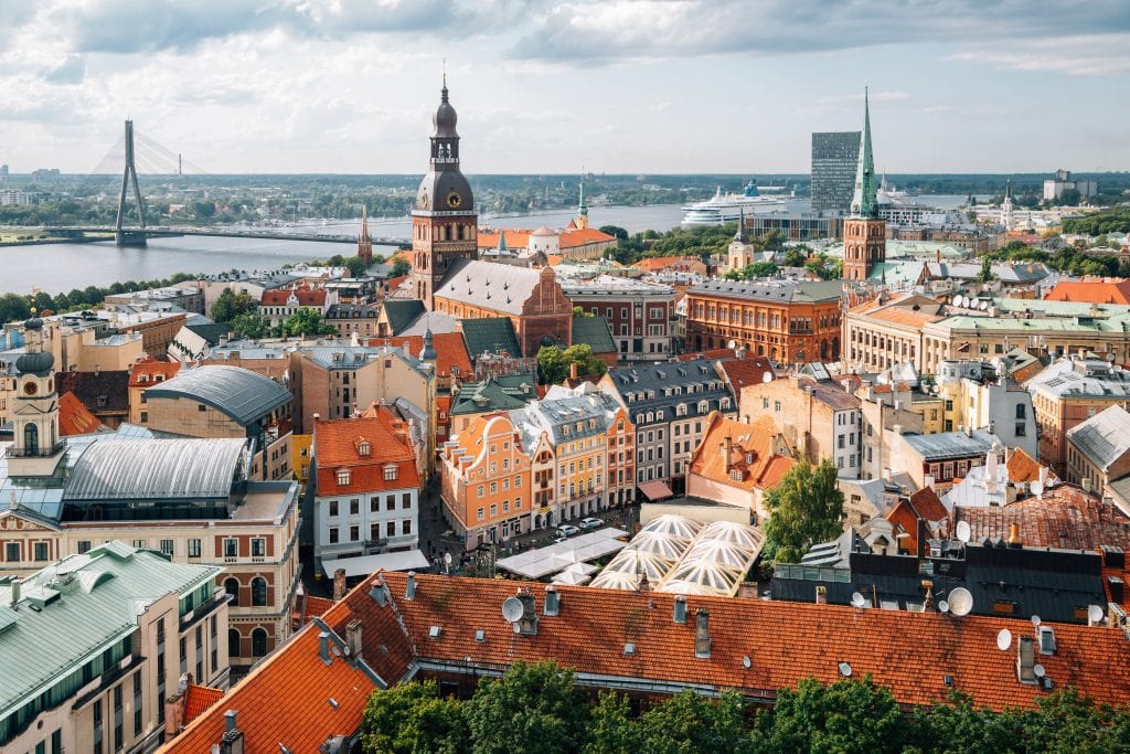 A skyline view of Riga: small Art Nouveau buildings in the foreground, a church tower and a bridge in the background. In the back is a river, and there seems to be a large cruise ship on it.