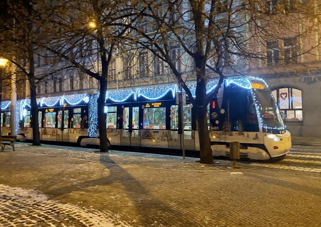 A tram on a snowy cobblestone stream in Prague. It's decorated with Christmas lights and images of Christmas trees and snowflakes painted on the side of it.