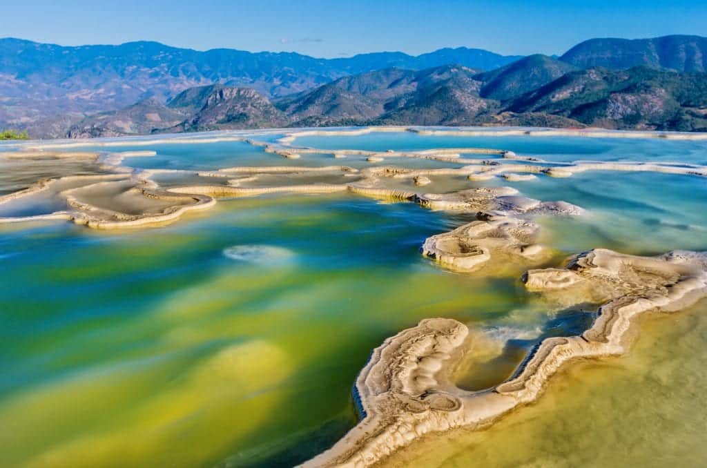 Almost alien-looking pools of water at the Hierve el Agua -- yellow fossilized petrified shapes surrounded by bright turquoise and teal pools. Mountains behind in the distance.