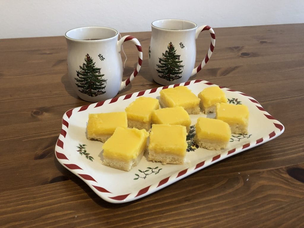 A Christmas-themed white platter with red and white striped edging topped with lemon squares, shortbread on the bottom and bright yellow curd on top. Behind the platter are two matching mugs with Christmas trees on them and red and white striped handles.