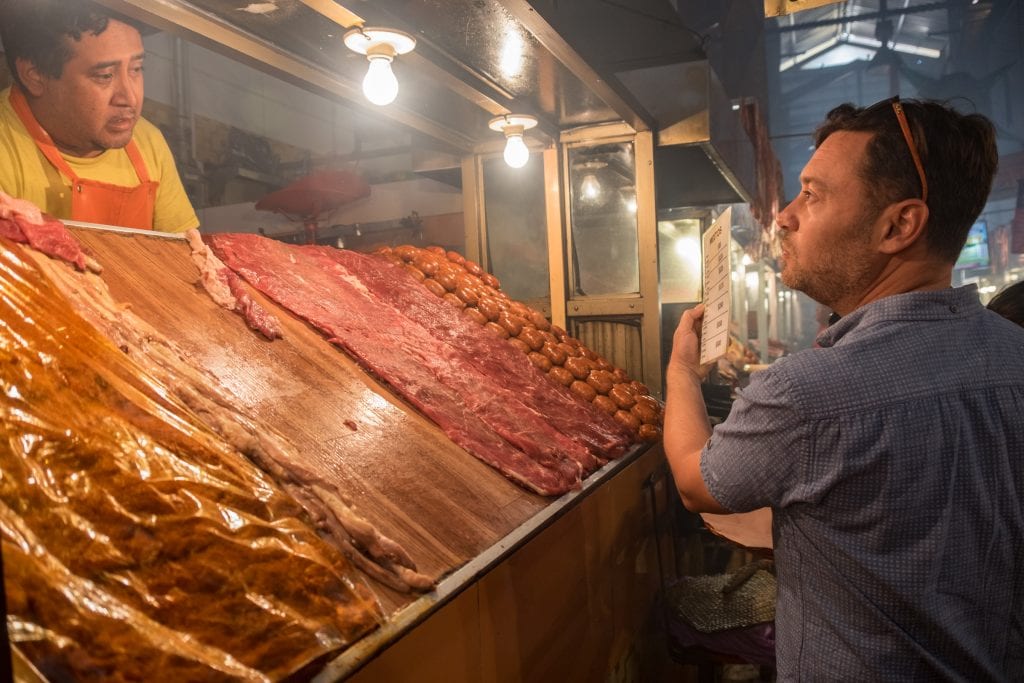 Charlie holds up a menu and talks to a butcher behind a wooden counter topped with lots of raw meat at Mercado 20 Noviembre.