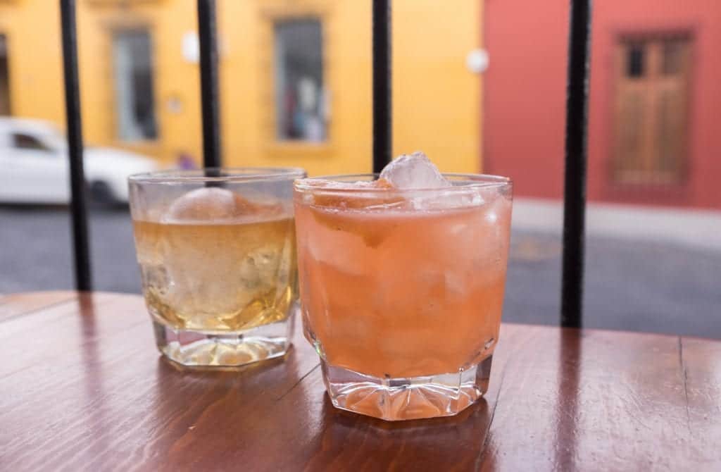 Two cocktails on a wooden shelf: one pale yellow and one pinkish orange, filled with large ice cubes.