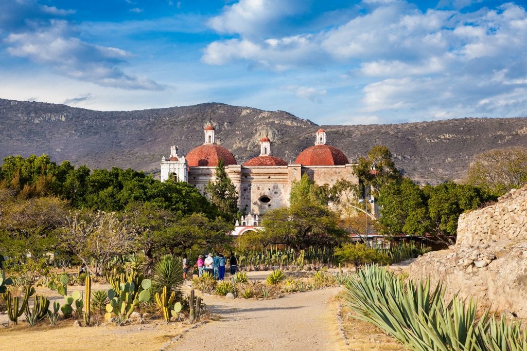 The town of Mitla with its beige stone building with three red domes. In front of it are cactus gardens and green trees; behind it are mountains.