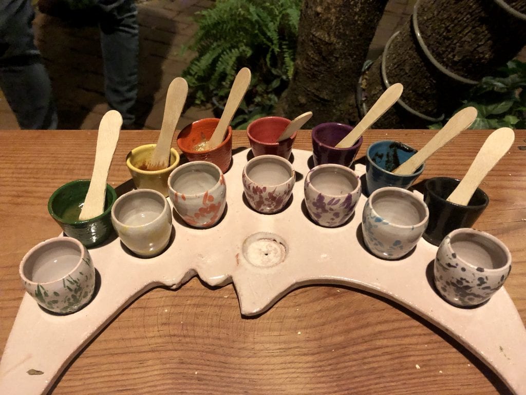 A white crescent moon-shaped plate topped with a row of seven white shot glass-sized glasses filled with mezcal, and a row of seven shot glasses painted the same colors as their moles within with wooden tasters sticking out.