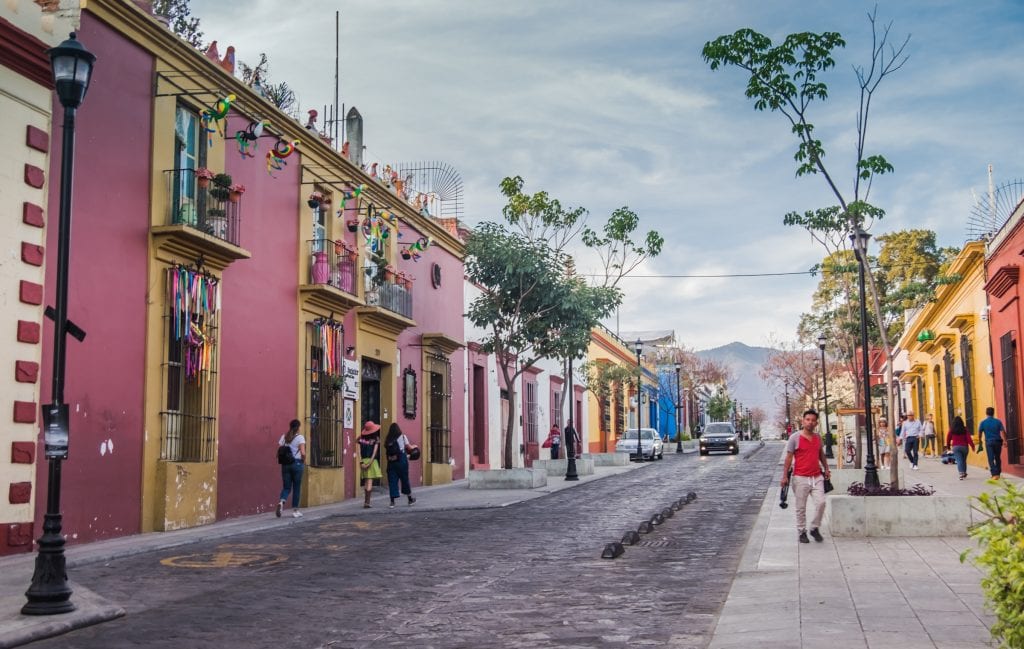 A bright and colorful street in Oaxaca with purple, yellow, blue, and white buildings in a row. There are trees and you see people walking down the street and mountains rising in the background.